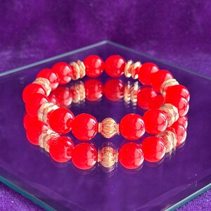 May include: A stretch bracelet featuring smooth, round, vibrant red beads alternating with smaller, textured, metallic gold-toned spacer beads. The bracelet is arranged in a circular shape, showcasing a consistent pattern of red and gold elements.