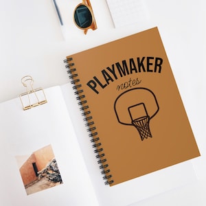 Puede incluir: Un cuaderno de espiral marrón con la inscripción "PLAYMAKER notes" y una ilustración de una canasta de baloncesto en negro. El cuaderno está abierto, mostrando páginas blancas, sobre una superficie blanca.