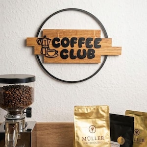 Könnte beinhalten: Eine Kaffee-Themenausstellung mit einer Kaffeemühle aus Edelstahl, gefüllt mit Bohnen, Kaffeepackungen und einer Tasse Kaffee. Ein Holzschild mit der Aufschrift "COFFEE CLUB" ist an der Wand angebracht und von einem schwarzen Metallring umgeben.