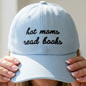 May include: Light blue baseball cap with the embroidered text "hot moms read books" in black. The cap has a curved brim and a button on top. A casual accessory for book lovers.
