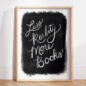 May include: A framed print featuring the words "Less Reality More Books" in white script on a black backdrop. The print is in a light wood frame, set against a white wall and a wooden floor.