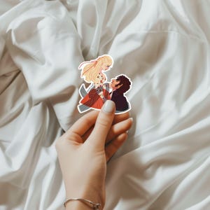 May include: A sticker featuring an illustration of a couple in a romantic embrace. The woman has long blonde hair and wears a red and floral dress. The man is in a black suit. The sticker is held by a hand.