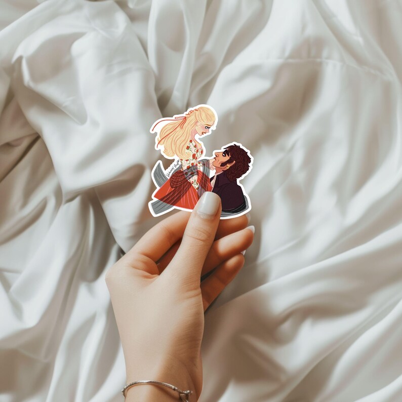 May include: A sticker featuring an illustration of a couple in a romantic embrace. The woman has long blonde hair and wears a red and floral dress. The man is in a black suit. The sticker is held by a hand.