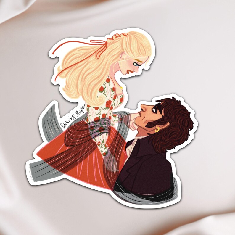 May include: A sticker featuring an illustration of a man and woman in a romantic embrace. The woman has long blonde hair and wears a dress with red roses. The man has dark hair and a black coat. The sticker has a white border.