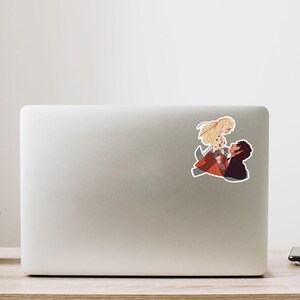 May include: A sticker featuring an anime-style illustration of a couple in a romantic embrace. The woman has long blonde hair and wears a red and orange dress. The man has dark hair and a black shirt. The sticker is placed on a laptop.