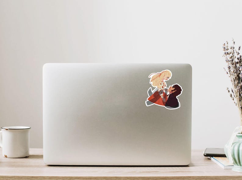 May include: A sticker featuring an anime-style illustration of a couple in a romantic embrace. The woman has long blonde hair and wears a red and orange dress. The man has dark hair and a black shirt. The sticker is placed on a laptop.