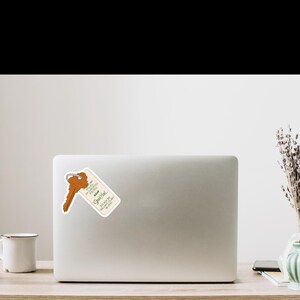 May include: A sticker featuring a vintage hotel key and tag. The key is brown, and the tag is cream-colored with green text that reads "Good Hotel" and other phrases. The sticker is placed on a white laptop.