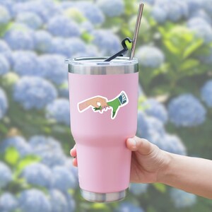 May include: A pink stainless steel tumbler with a sticker of two hands reaching out to touch each other. One hand is human and the other is green.