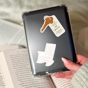 May include: A clear phone case with three stickers: a key tag that reads "Grand Hotel", a key, and a rocking chair. The key tag has the text "WE GUARANTEE POSTAGE". The phone case is held over an open book.