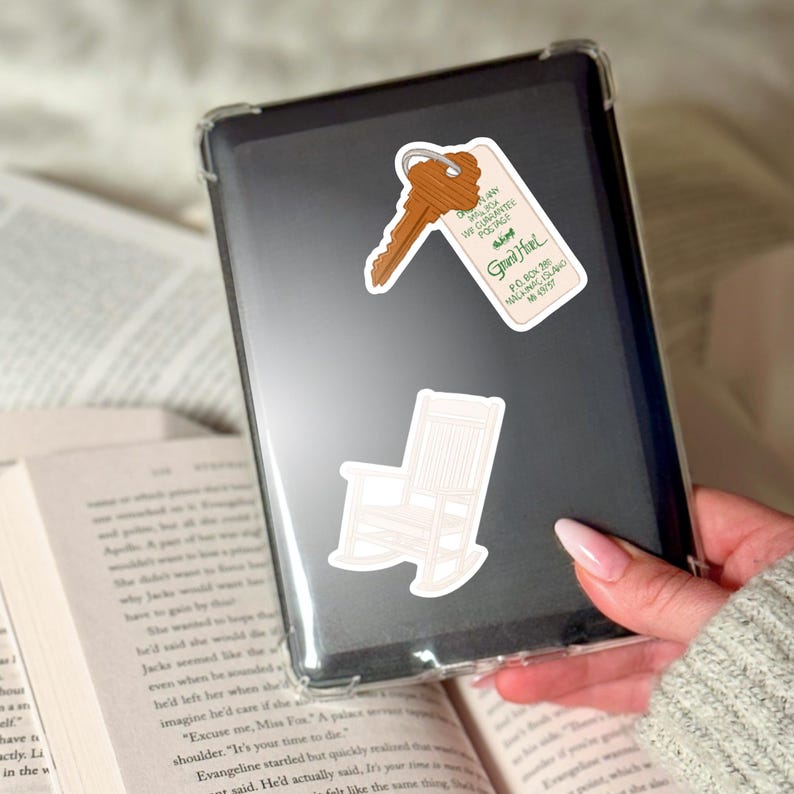 May include: A clear phone case with three stickers: a key tag that reads "Grand Hotel", a key, and a rocking chair. The key tag has the text "WE GUARANTEE POSTAGE". The phone case is held over an open book.