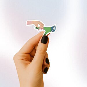May include: A sticker featuring a green hand reaching out to a pink hand with long pink nails. The sticker is on a white background.