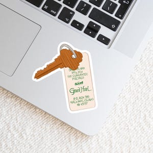 May include: A sticker featuring a vintage-style key and hotel tag. The key is brown, and the tag has the text "Grand Hotel" in green, along with address details. The sticker is placed on a laptop.
