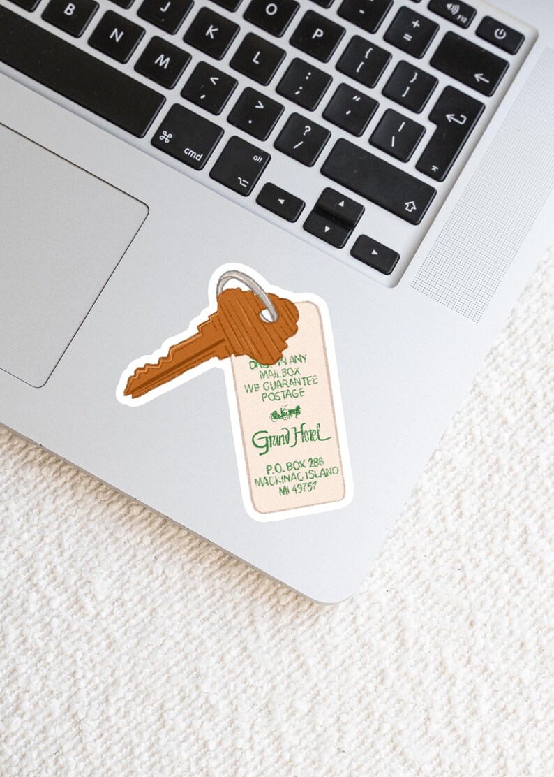 May include: A sticker featuring a vintage-style key and hotel tag. The key is brown, and the tag has the text "Grand Hotel" in green, along with address details. The sticker is placed on a laptop.