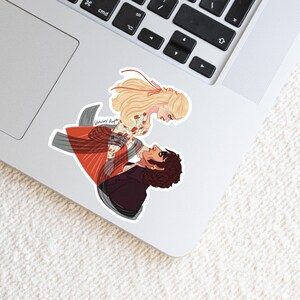 May include: A sticker featuring an illustration of a man and woman in a romantic embrace. The woman has long blonde hair and a floral dress. The man wears a dark suit. The sticker is on a laptop.
