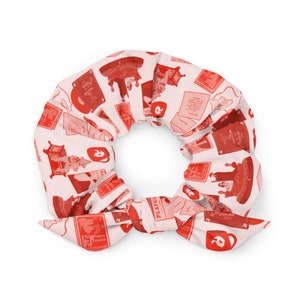 May include: A white scrunchie with a red pattern featuring opera-themed illustrations, including playbills, stage scenes, and the Phantom of the Opera logo. The scrunchie has a bow detail.