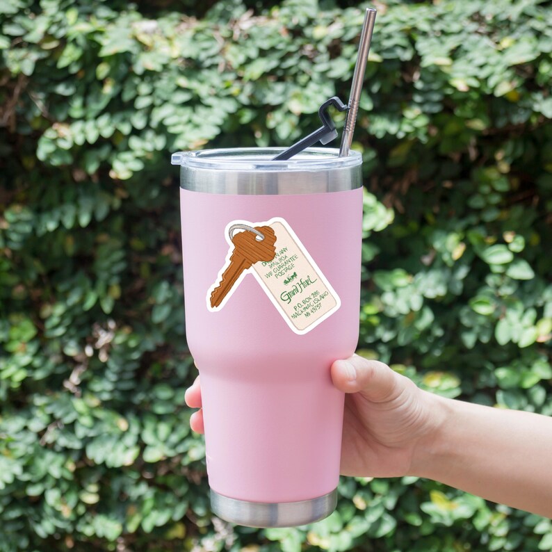 May include: A light pink insulated tumbler with a silver rim and lid. A sticker of a brown key and a tag that reads "Grand Hotel" is affixed to the tumbler. A stainless steel straw and black straw tip are inserted into the lid.