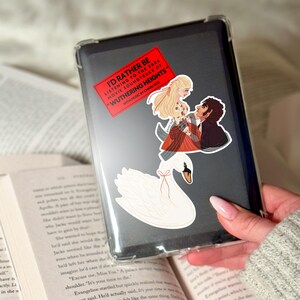 May include: A clear phone case with stickers. The stickers include a white swan, and a graphic of two people with the text "I'D RATHER BE LISTENING TO THE 2026 MOVIE SOUNDTRACK OF 'WUTHERING HEIGHTS' WITH MUSIC BY CHARLI XCX" on a red rectangle.