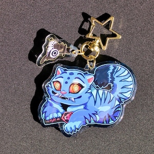May include: A blue cat-like character keychain with orange eyes and a red gem. The keychain has a gold star-shaped clasp and a smaller charm with a black and white design. The cat has blue stripes and is lying down.