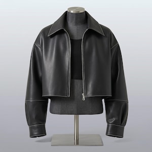 Könnte beinhalten: Schwarze Lederjacke mit kurzem Design, Kragen, langen Ärmeln und silbernem Reißverschluss. Die Jacke wird auf einer Schaufensterpuppe präsentiert, die ihre maßgeschneiderte Passform und moderne Ästhetik zeigt. Die Nähte sind weiß.