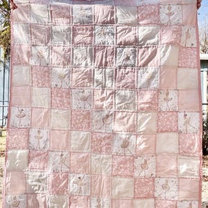 May include: A patchwork quilt in shades of pink and white, featuring a repeating pattern of squares. Some squares have a floral design, while others depict a ballerina. The quilt is held up, showcasing its size and design. The edges are finished with a pink border.