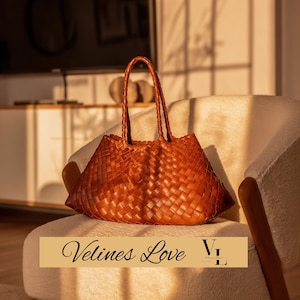 May include: A woven brown leather tote bag with braided handles sits on a cream-colored armchair. The bag has a trapezoid shape and a textured surface. The text "Velines Love" is displayed on a banner at the bottom.