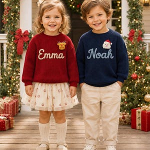 Può includere: Un maglione in maglia rosso con il nome Emma e una toppa a forma di renna, abbinato a una gonna in tulle color crema, e un maglione in maglia blu navy con il nome Noah e una toppa di Babbo Natale, abbinato a pantaloni beige e scarpe da ginnastica bianche.