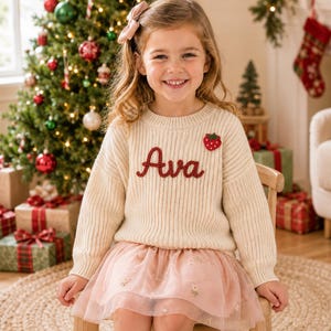 May include: A cream-colored ribbed knit sweater featuring the name Ava embroidered in dark red yarn across the chest, accented by a small red strawberry patch. The sweater is paired with a light pink layered tulle skirt with floral embroidery.