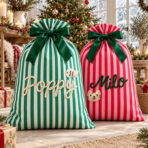 May include: Two fabric gift bags with vertical stripes, one green and white and one red and pink, each topped with a dark green satin ribbon bow. The green bag features the name Poppy in white script and a small fish patch, while the red bag displays the name Milo in green script and a small cat face patch.