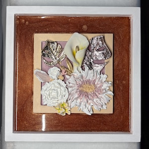 Könnte beinhalten: Quadratisch gerahmtes Blumenkunstwerk. Das Kunstwerk hat einen weißen Rahmen, der eine braune und helle Holzumrandung umschließt. Im Inneren sind verschiedene geformte Blumen in Weiß, Rosa und Gold auf einem lavendelfarbenen Hintergrund angeordnet.