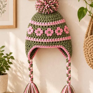 Puede incluir: Un gorro de ganchillo color verde oliva con un gran pompón en la parte superior, una banda de motivos florales rosas y dos largas orejeras trenzadas que terminan en gruesas borlas multicolores de hilo verde oliva, rosa y blanco.