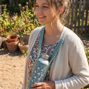 May include: A light blue crocheted water bottle holder with a strap, holding a silver and white water bottle. The holder has a geometric design. The person is wearing a floral top and a cream cardigan.