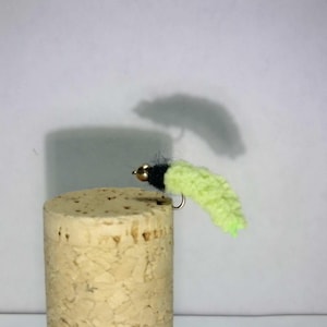 May include: A handmade fishing fly with a black head and a chartreuse body. The fly is attached to a small hook and is resting on a cork. The fly is designed to attract fish.