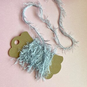 May include: A close-up shot of a light blue, fluffy yarn strand and a brown paper tag with a scalloped edge. The yarn is draped over the tag and has a soft, feathery texture. The background is a solid pink color.