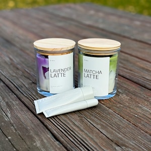 Puede incluir: Dos frascos de vidrio con tapas de madera, etiquetados como "Lavender Latte" y "Matcha Latte", están sobre una superficie de madera. El frasco de lavanda contiene cera morada, y el frasco de matcha contiene cera verde. Dos tubos blancos están frente a los frascos.