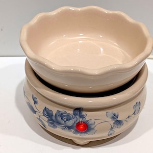 May include: A two-piece beige ceramic wax warmer with a floral design. The top dish has a scalloped edge and sits atop a base decorated with blue flowers and leaves. A red light is visible on the front of the base.