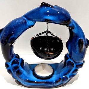 May include: A ceramic candle holder featuring two blue dolphins forming a circle. A black hanging bowl is suspended in the center, and a white candle sits at the base. The dolphins have a glossy finish.