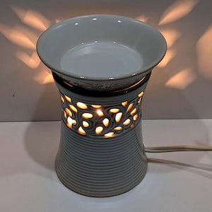 May include: A gray ceramic essential oil warmer with a round top and a decorative floral pattern around the middle. The warmer is lit from within, casting a warm glow and creating interesting shadows. A power cord extends from the base.