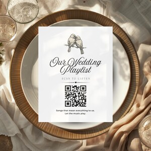 May include: A white card with the text "Our Wedding Playlist" and a QR code, placed on a white plate with a gold rim. The card features a drawing of two birds. The setting includes glasses and floral arrangements.
