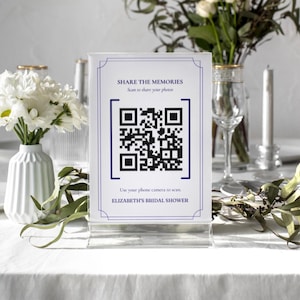 May include: A clear acrylic sign with a QR code and text that reads "SHARE THE MEMORIES. Scan to share your photos. Use your phone camera to scan. ELIZABETH'S BRIDAL SHOWER." The sign is on a table with white flowers and greenery.