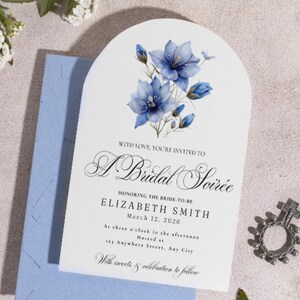 May include: An arched invitation card with blue floral accents and the text "A Bridal Soirée." The card includes details for Elizabeth Smith's bridal event on March 12, 2026. A small, decorative silver ring is also visible.