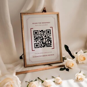 May include: A wooden framed sign with the text "SHARE THE MEMORIES" and a QR code. The sign is displayed on a white surface with white roses. The text "ELIZABETH'S BRIDAL SHOWER" is at the bottom of the sign.
