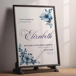 May include: A framed sign with a white background and blue floral accents. The sign reads "LET'S CELEBRATE The Bride-To-Be Elizabeth WELCOME TO THE BRIDAL SHOWER We're so glad you're here 04.23.2026 The Grand Garden California, USA."