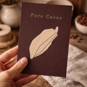 Może przedstawiać: Ciemnobrązowa karta z napisem "Pure Cocoa" u góry i beżową ilustracją strąka kakaowca. Karta trzymana jest w dłoni, na drewnianej powierzchni i lnianej tkaninie w tle.
