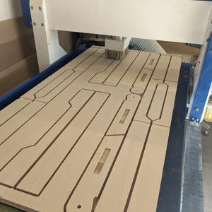 May include: A CNC router machine with a blue and white frame is cutting multiple elongated wooden components from a large flat sheet of light-colored plywood. The machine features a central spindle with a brush attachment and a black cable track.