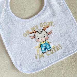May include: A white terry cloth bib featuring a central illustration of a cartoon goat wearing blue overalls and a yellow shirt. The text "OH MY GOAT, I'M CUTE!" is printed in gold lettering above and below the goat character on the fabric.