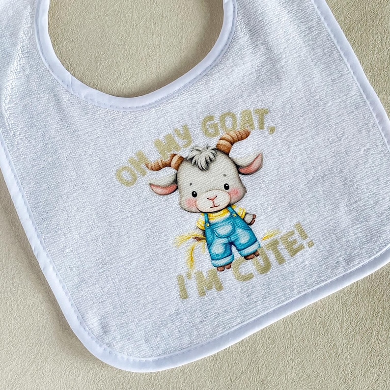 May include: A white terry cloth bib featuring a central illustration of a cartoon goat wearing blue overalls and a yellow shirt. The text "OH MY GOAT, I'M CUTE!" is printed in gold lettering above and below the goat character on the fabric.