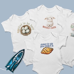 May include: Three white baby bodysuits featuring graphic prints. One shows a bird nest with eggs and the text "eggcited to be here," another a lamb with "WOOLY CUTE," and the third a croissant with "flake it till you make it." A blue iron is included.