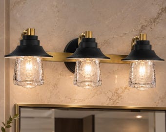 May include: A three-light vanity fixture featuring matte black metal shades and clear seeded glass covers. The lights are mounted on a horizontal gold-toned metal bar with matching gold-toned hardware connecting each shade to the base.