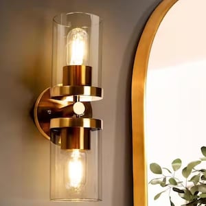 May include: A wall-mounted light fixture featuring two cylindrical clear glass shades held by a brushed gold metal frame. The design includes two vertically stacked light bulbs that emit a warm glow, connected by a central circular gold accent.