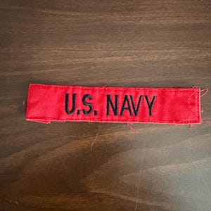 May include: A red fabric patch with the text "U.S. NAVY" embroidered in black. The patch is rectangular and appears to be a uniform accessory.